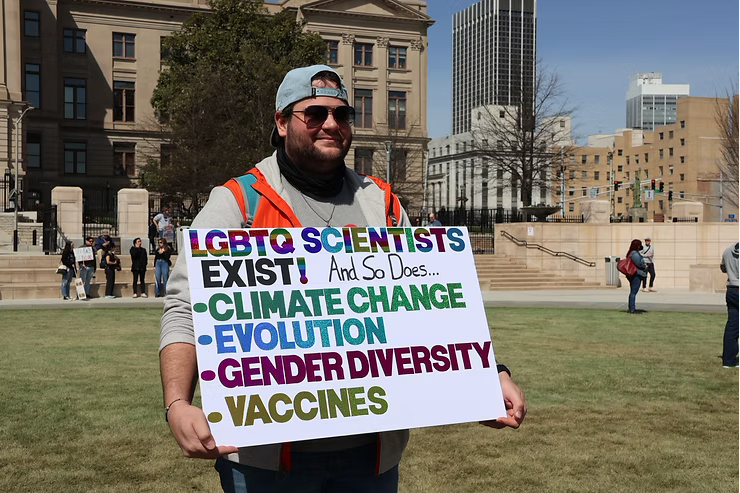 A protester on the lawn outside the Georgia State Capital holding a sign that reads "LGBTQ Scientists exists! and so does climate change, evolution, gender diversity, vaccines"